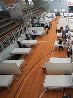 The photo shows a large gymnasium with lots of beds in rows. 