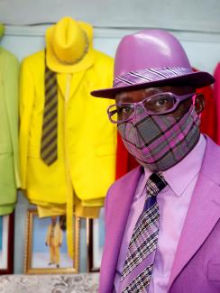 Picture shows James smartly dressed in a well-appointed purple suit with a matching mask. In the background are striking blue and orange and green suits hanging on the wall. 