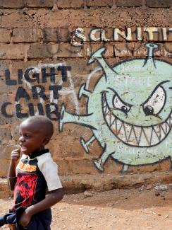 This is a powerful photo of a young boy walking in front of a sinister depiction of the virus with a face that appears to be sneeringly looking at the boy. 