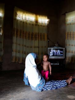 The photo shows mother and child on the floor of a large, spare living room darkened with curtains and illuminated by a TV in the corner. 