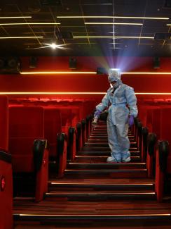 The photo is striking, showing a worker spraying disinfectant wearing a blue suit descending down a theater staircase where all the seats and carpet are bright red. 