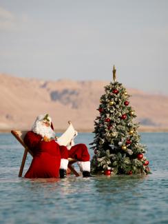 A man, wearing a red Santa Claus costume complete with a hat, beard, and bag full of toys, holds a scroll as he poses for a picture while sitting next to a decorated Christmas tree on a salt formation in the Dead Sea, near Ein Bokeq, Israel on November 15, 2020.