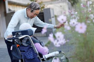 An elderly man helps his wife in a wheel chair to enjoy some pink flowers