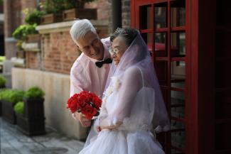 An elderly couple in wedding attire pose with a bouquet of red flowers