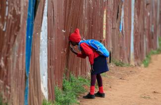 A girl in a red school uniform peers through a school yard fence in Kenya