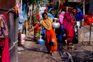 A woman in an orange dress carries a water jar after collecting water from a common tap. In the background are other women in colorful garments who don't have direct access to water at home in Dhaka, Bangladesh, February 21, 2022. 