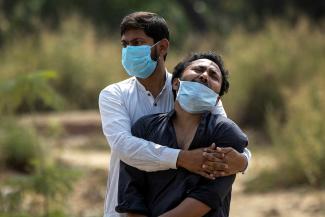 a young man wearing a white linen shirt and a blue surgical mask consoles his relative who's crying 