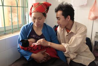 A mother in a blue shirt and orange head wrap breastfeeds her newborn as she sits next to her partner, who wears a white collared shirt as he fawns over the child. The two sit in front of a window with green metalwork in a well-lit white hospital room.