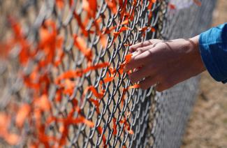 A white hand with the cuff of a blue jacket reaches out to tie an orange ribbon onto a chain-link fence covered in orange ribbons in honor of those killed by gun violence