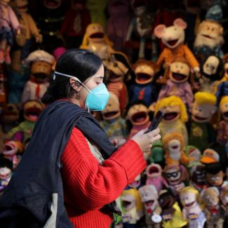 A woman in a red coat and blue mask checks her phone as she walks by a stall with colorful children's puppets