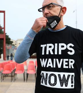 A man adjusts his face mask while wearing a black t-shirt that reads "TRIPS WAIVER NOW!" He is standing in front of rows of empty chairs.