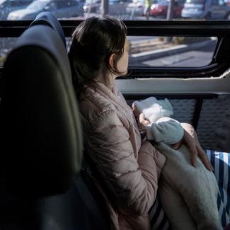 A Ukrainian refugee mother in a pink coat breastfeeds her baby while looking out the window of a train.