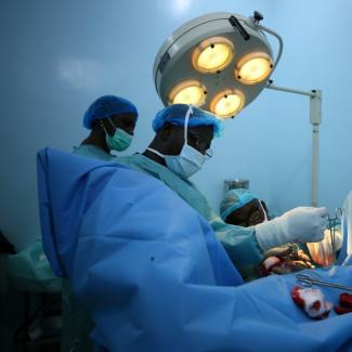 Nigerian surgeons in blue gowns and masks operate on a patient hidden beneath a blue sheet under a large surgical lamp in a room with blue walls. 