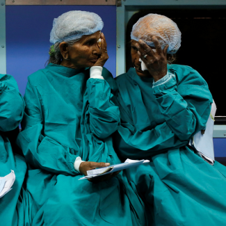 Two patients dressed in green medical gowns and plastics caps sit on a bench and cover their eyes as they wait for cataract surgery on the Lifeline Express, a hospital built inside a seven-coach train, at a railway station in Jalore, India, on March 31, 2018. 