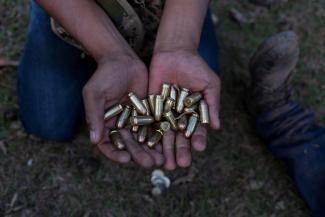 A community police officer in blue jeans squats low to the ground and holds out two hands full of shiny metal 45mm caliber bullets