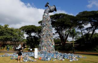 At UN Headquarters in Nairobi, Kenya, a female delegate wearing a long-sleeved black dress and a blue surgical mask looks at a 30-foot sculpture made out of plastic bottles that resembles a faucet, outside the UN Environment Programme Headquarters, in Nairobi, Kenya 