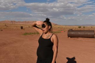 A pregnant woman in a black tank top raises her hand to shield her eyes from the sun 