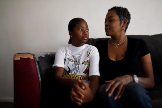 A young Black girl in a white t-shirt with a ponytail sits next to her mother who has short hair and is wearing a black t-shirt on a tattered red and leather couch in front of a white wall