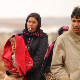 An Syrian woman with a serious expression on her face dressed in dark colors holds a child who is partially obscured by a bright orange scarf draped over his head. On her right , a young man in a beige winter coat walks alongside her through a desert landscape. 