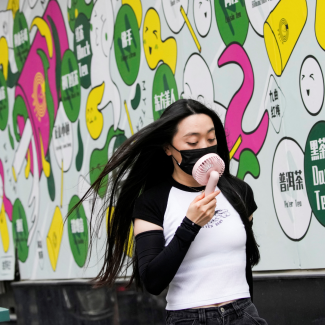 A woman in a white and black t-shirt wears a face mask uses a handheld fan as she walks on a street on a hot day, following the coronavirus disease (COVID-19) outbreak in Shanghai, China, on July 19, 2022. A colorful white, green, pink and yellow mural is pictured in the background.