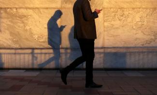 A man using a smart phone walks through London's Canary Wharf financial district in the warm yellow evening light, casting a shadow in London, United Kingdom, on September 28, 2018.