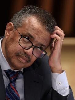 WHO Director-General Tedros Adhanom Ghebreyesus scratches his head with a quizzical expression while seated at a wooden podium 