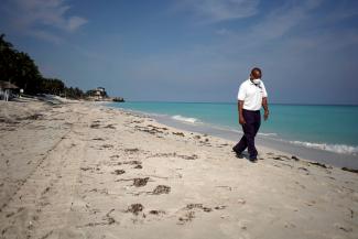 A security agent wearing a white protective mask, white polo shirt, and black slacks, walks along the shoreline of a white sand beach with crystal blue waters in Cuba
