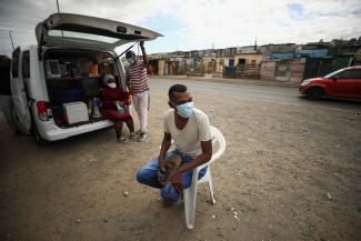 A young man wearing a white t-shirt, blue jeans, and a blue surgical face mask, sits on a plastic chair outside a mobile TB testing clinic.