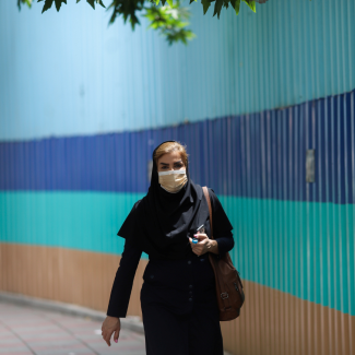 A woman walks along a street in Tehran, Iran, on May 26, 2021. Behind her stretches a wall with thick colorful horizontal stripes in shadess of blue and aqua.