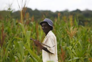 An older, African man wearing a black bucket hat and beige shirt, stands in a field. 