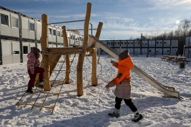 a child in an orange puffer jacket pushes their friend on a wooden swing in the snow